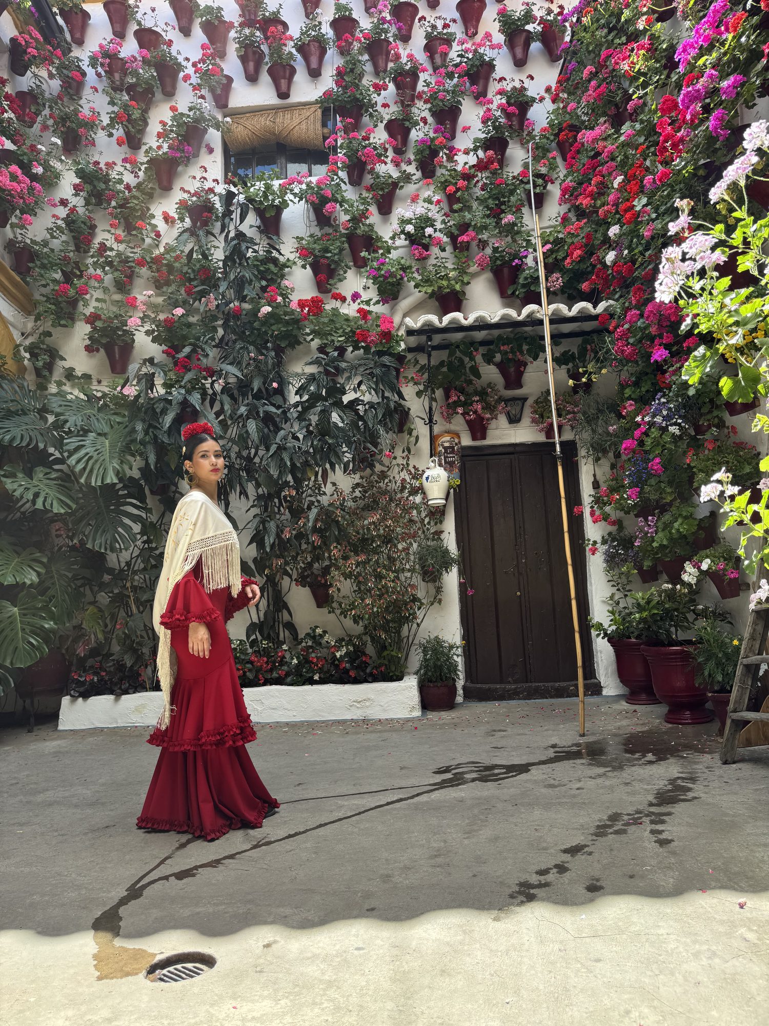 Bailaora con traje de flamenca ante una pared de patio cordobés cubierta de macetas de geranios